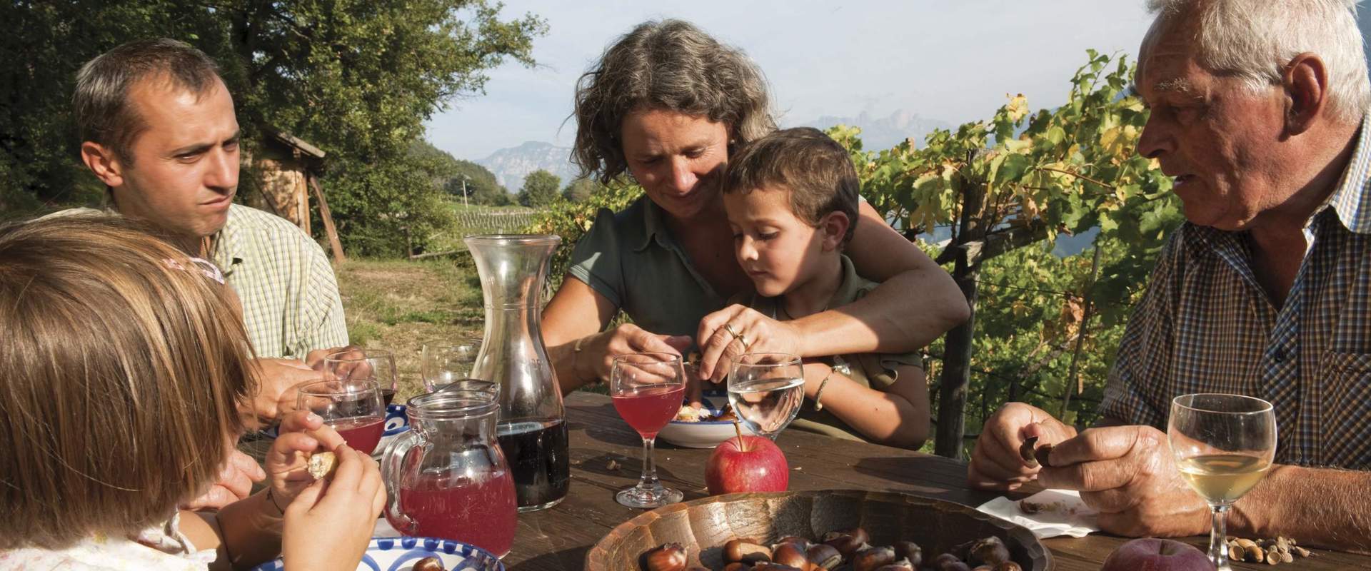 Famiglia seduta ad un tavolo all'aperto e mangia castagne e beve vino.