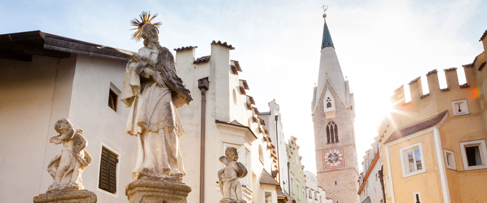 Centro storico di Bressanone. Statua di un santo affiancato da due angeli su un arco di pietra, alle sue spalle case di color pastello ed il campanile della città.
