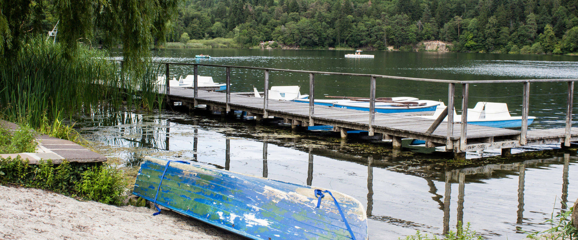 Una barca blu giace sulla riva sabbiosa del Lago di Monticolo. Sullo sfondo, la passerella con i pedalò azzurri conduce al centro del lago.