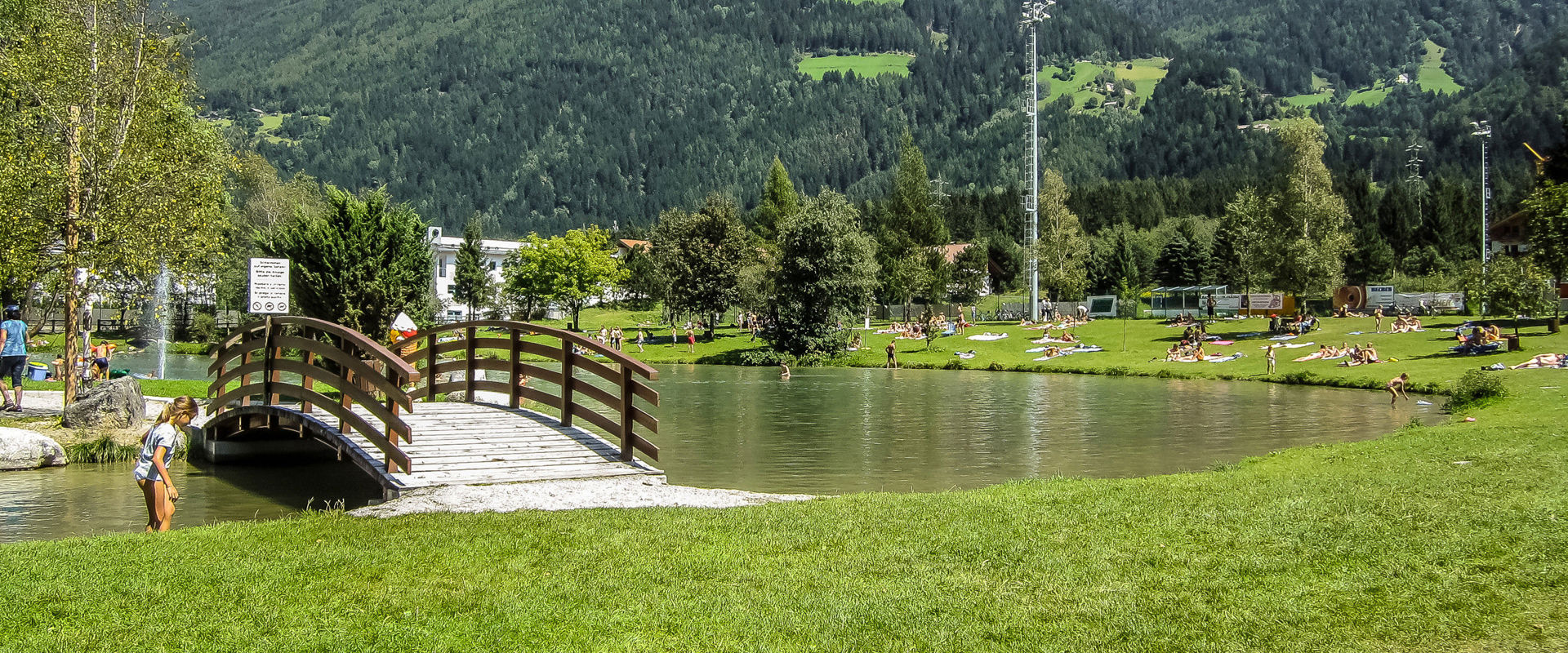 Romantico lago balneabile con un piccolo ponte, immerso nel verde e circondato da montagne.