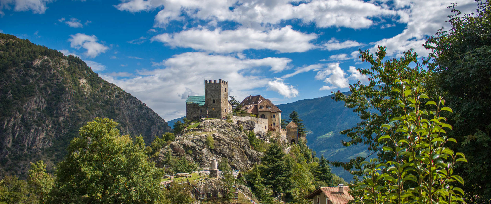 Castel Juval Il Castel Juval è arroccato su una collina rocciosa circondata da alberi, montagne, una casa e una piccola capanna.