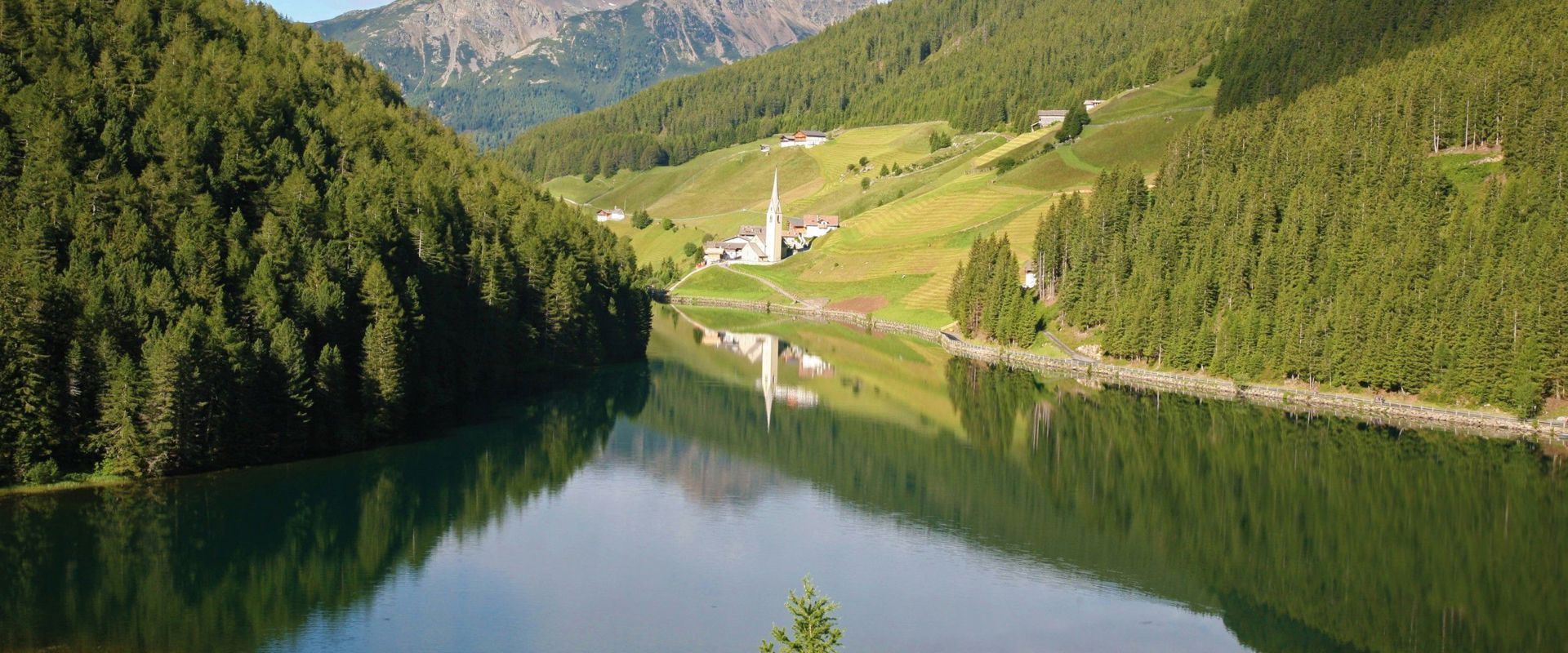 Lago in posizione fantastica con panorama montano e vista su una piccola chiesetta.