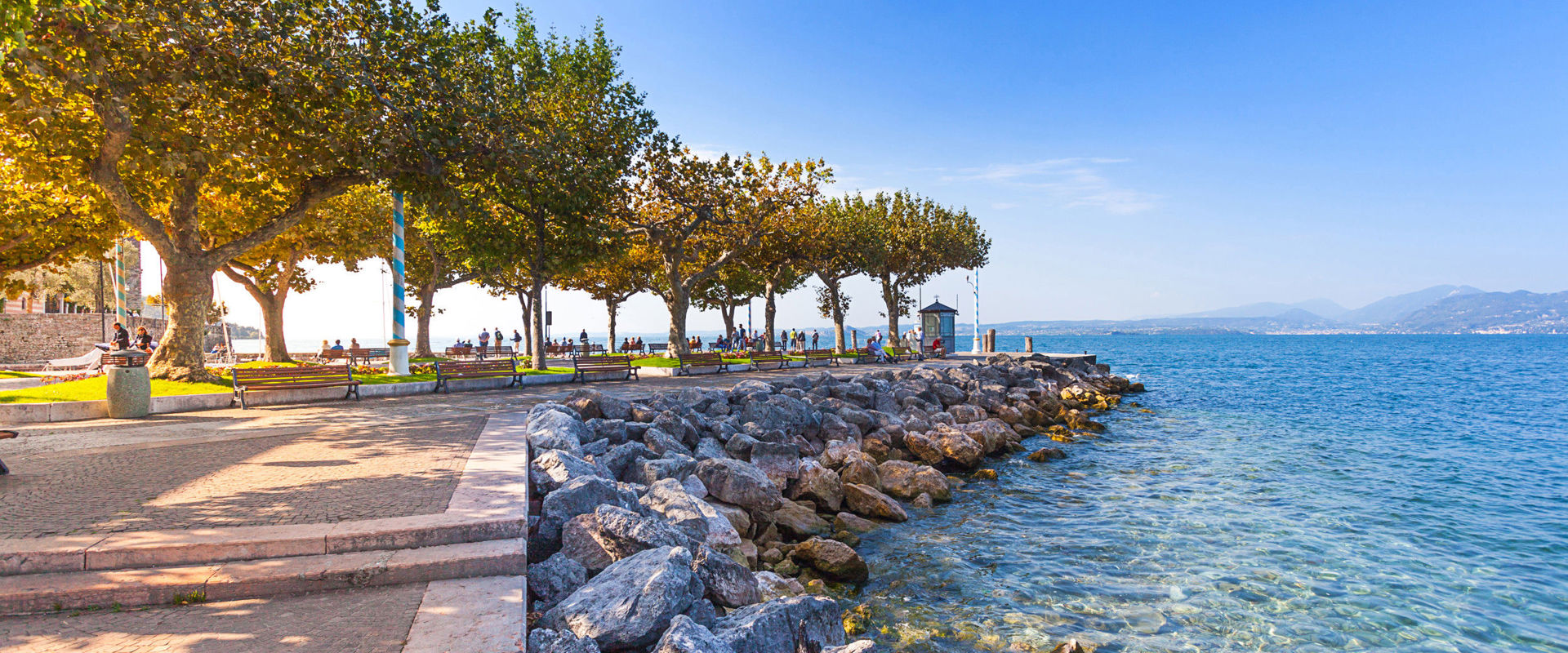 Passeggiata pavimentata con un viale di alberi a latifoglia lungo la riva del lago.