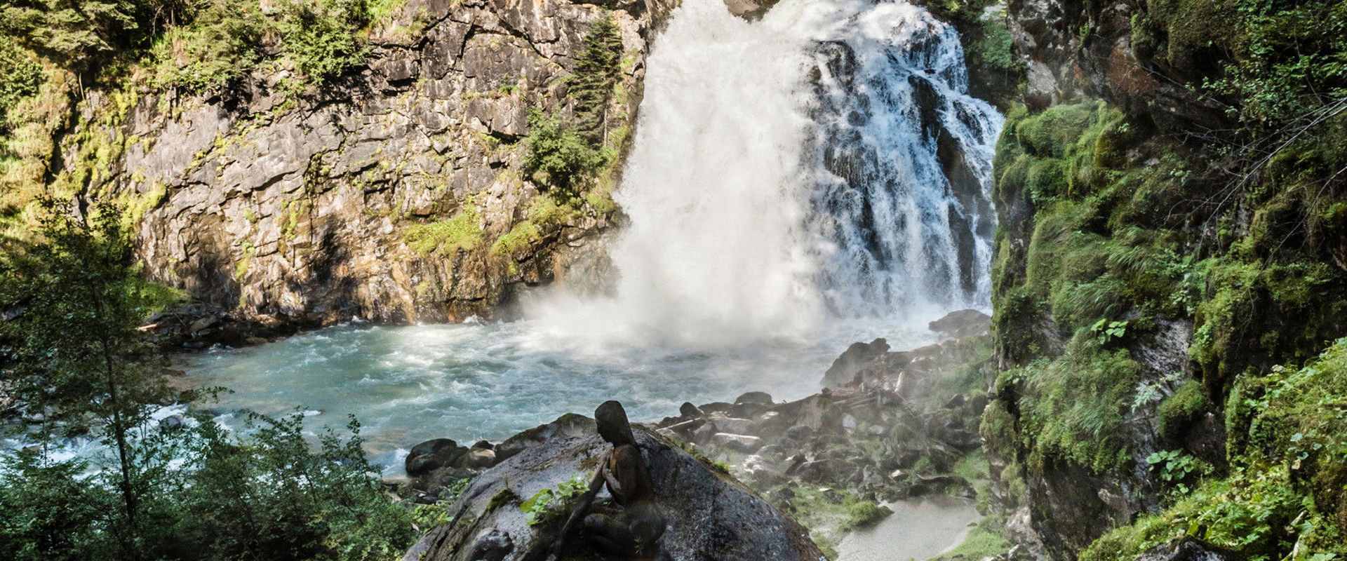 Cascate di Riva Cascata che scende sulle rocce. A sinistra e a destra dell'immagine si vedono ripide pareti rocciose con alte conifere.