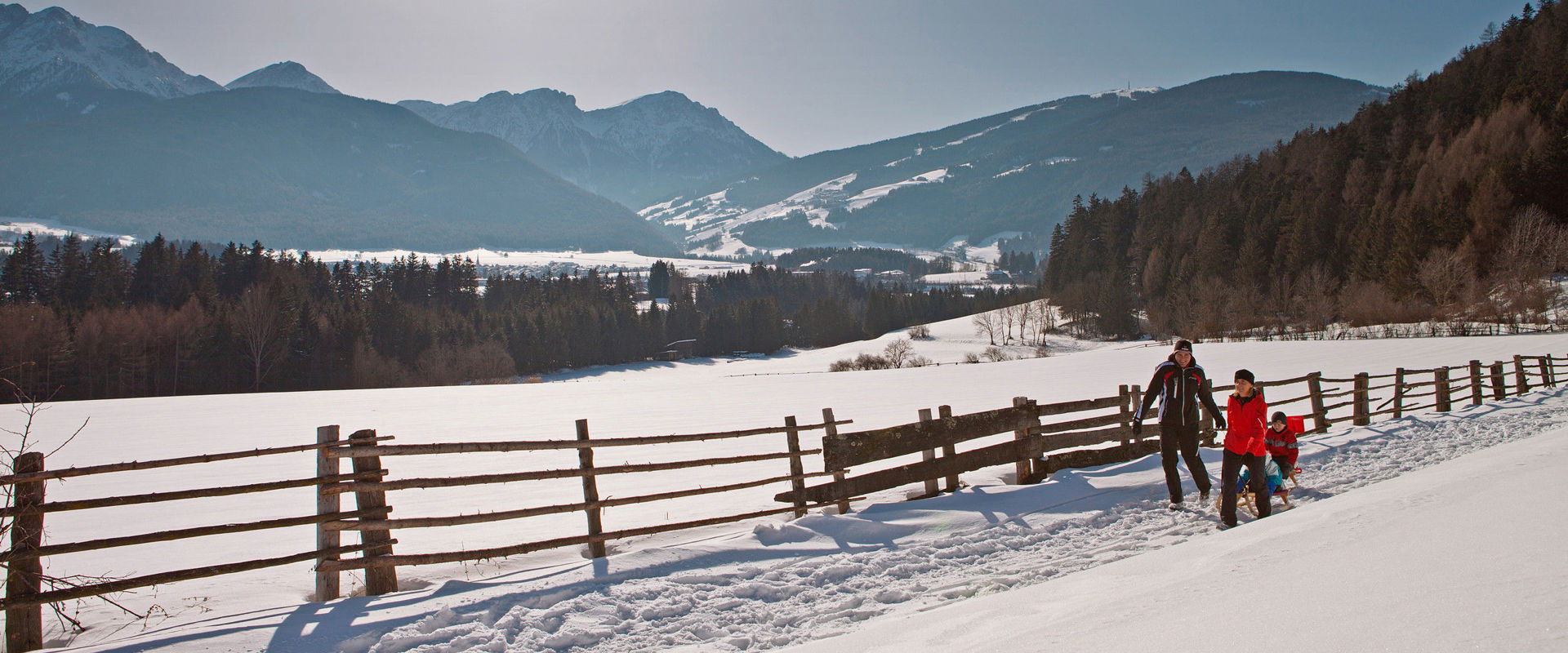 Inverno a Rasun-Anterselva. Uomo e donna che trainano due bambini su delle slitte in un soleggiato paesaggio invernale.