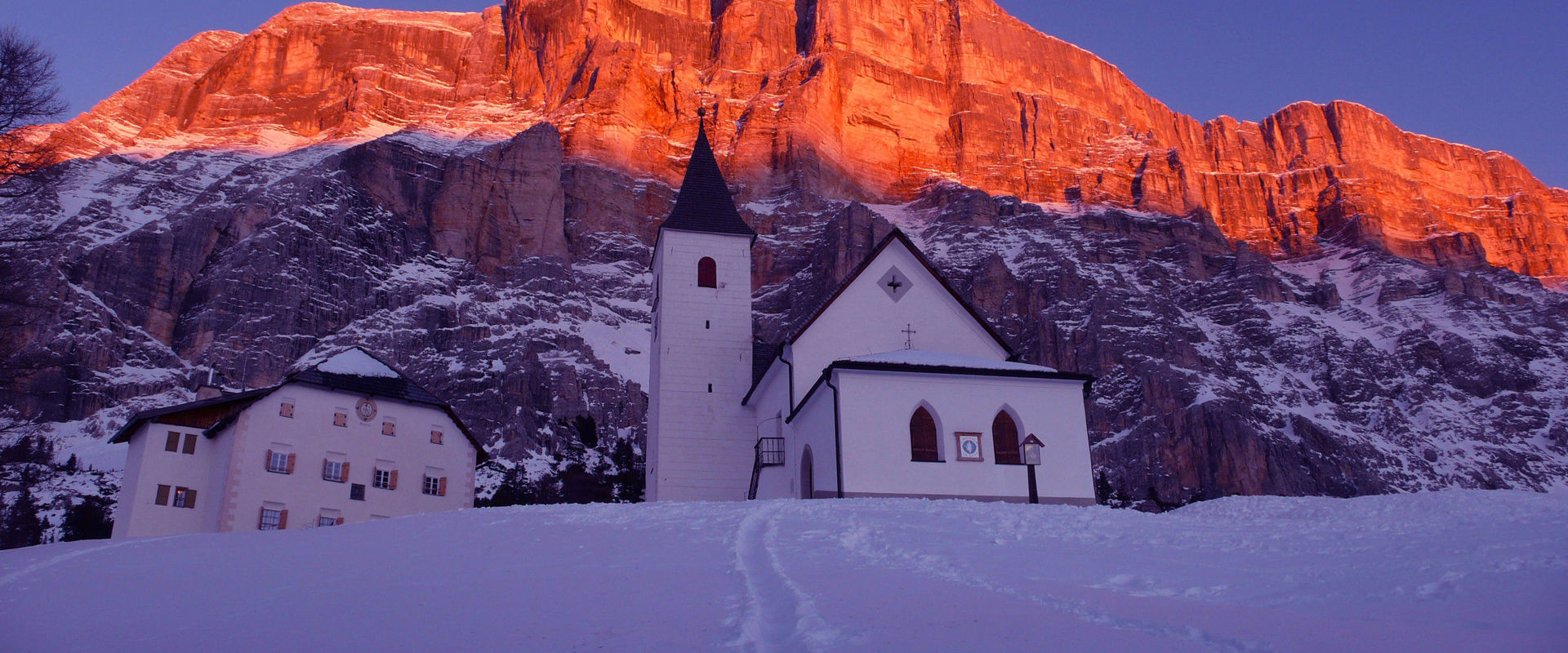 Pedraces Chiesetta di Santa Croce, meta di pellegrinaggi, con le Dolomiti colorate di rosso al tramonto sullo sfondo