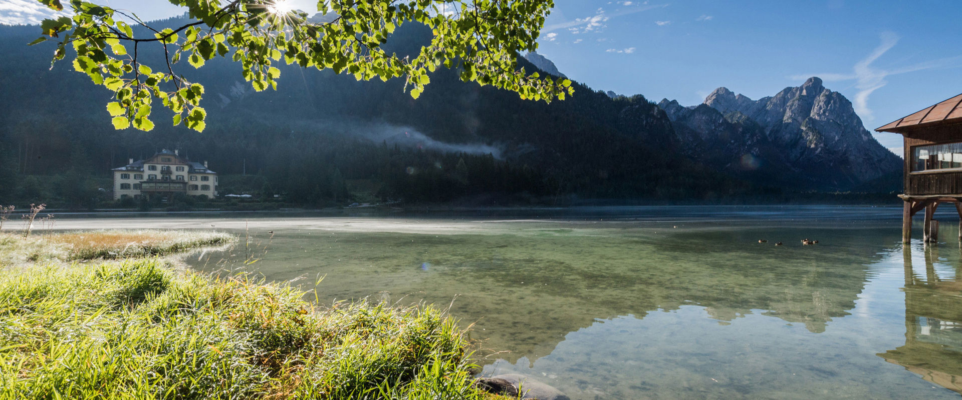 Riva di un lago limpido tra le montagne, sullo sfondo si intravedono degli edifici.