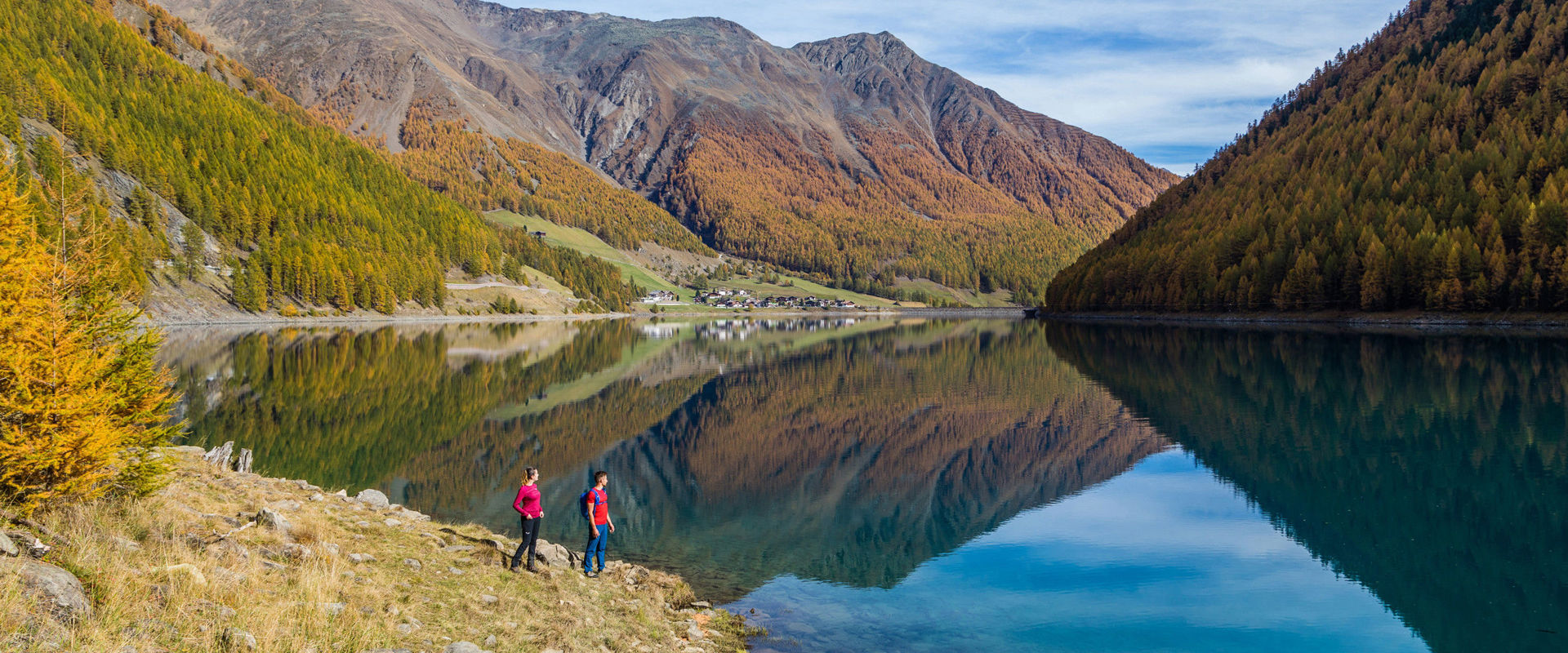 Giovane coppia in piedi sulla riva del lago di Vernago. Le montagne circostanti si riflettono nell'acqua.