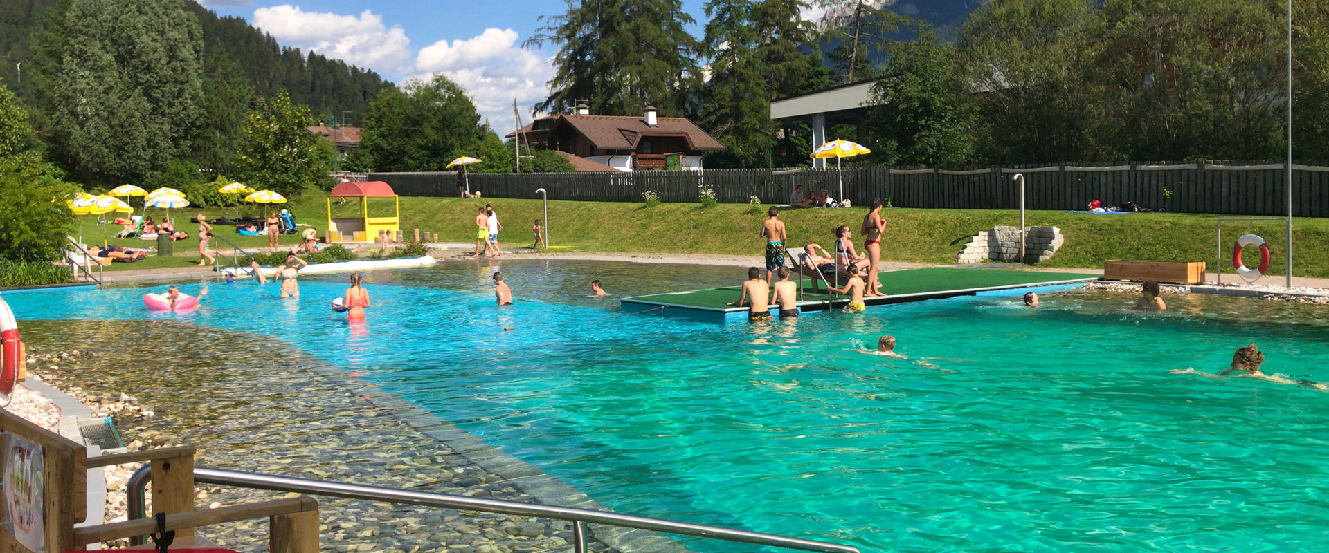 Lago balneabile con piscina per bambini, pontile, docce e prato per prendere il sole. Sullo sfondo si vedono case e alberi.