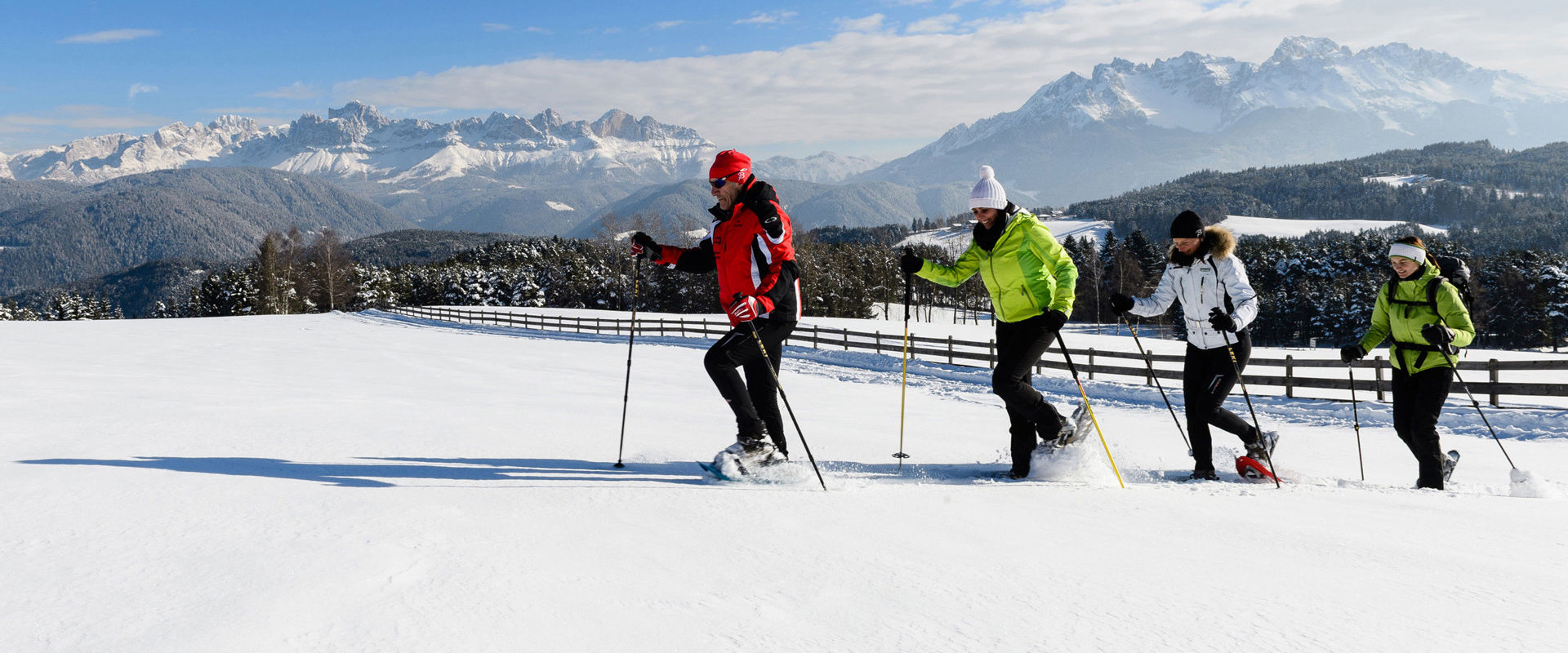 4 escursionisti con le ciaspole camminano nella neve alta su un prato recintato. Sullo sfondo si vede un paesaggio montano mozzafiato.