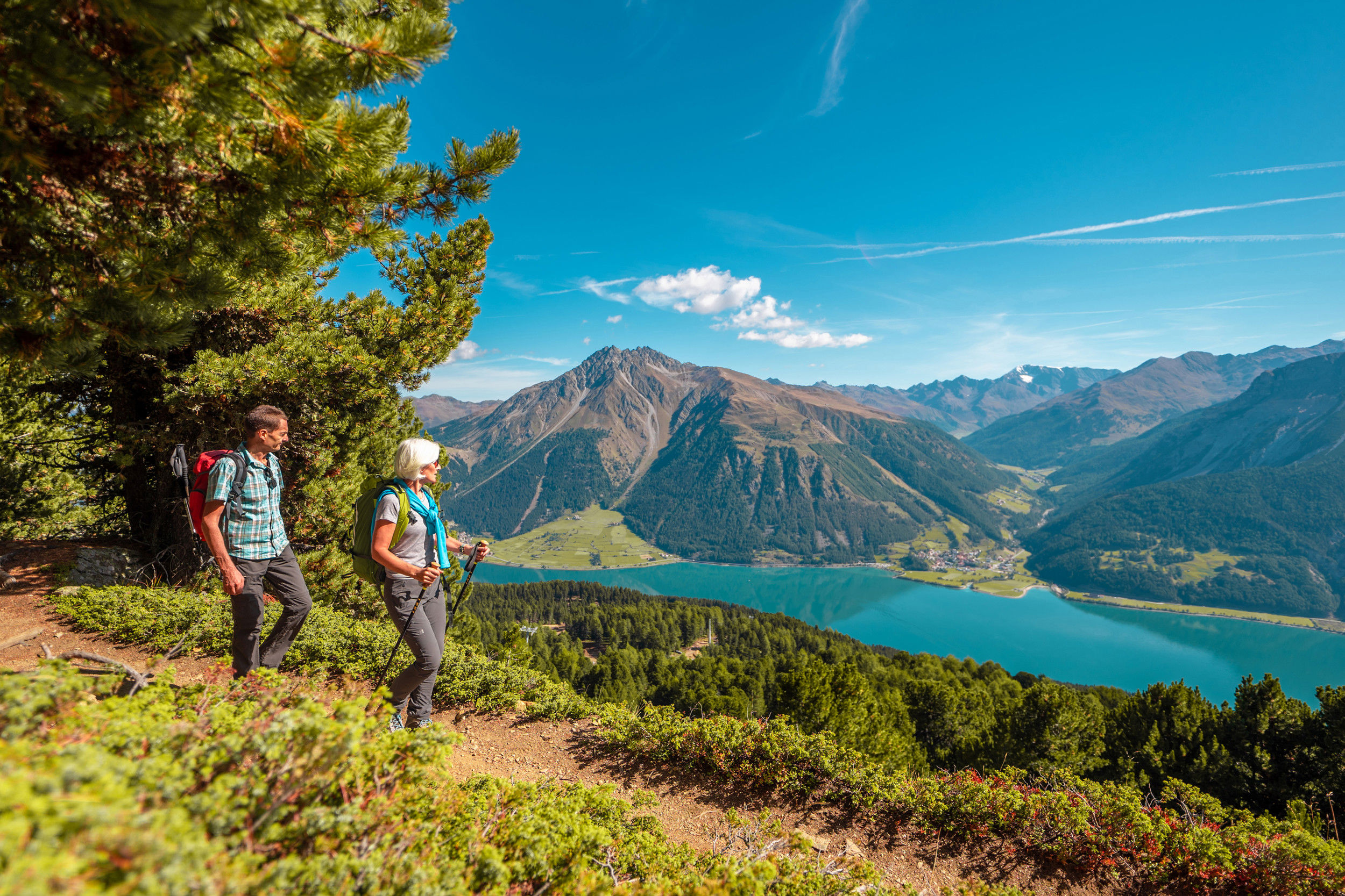Due escursionisti guardano il lago di Resia e il paesaggio montano dall'alto.