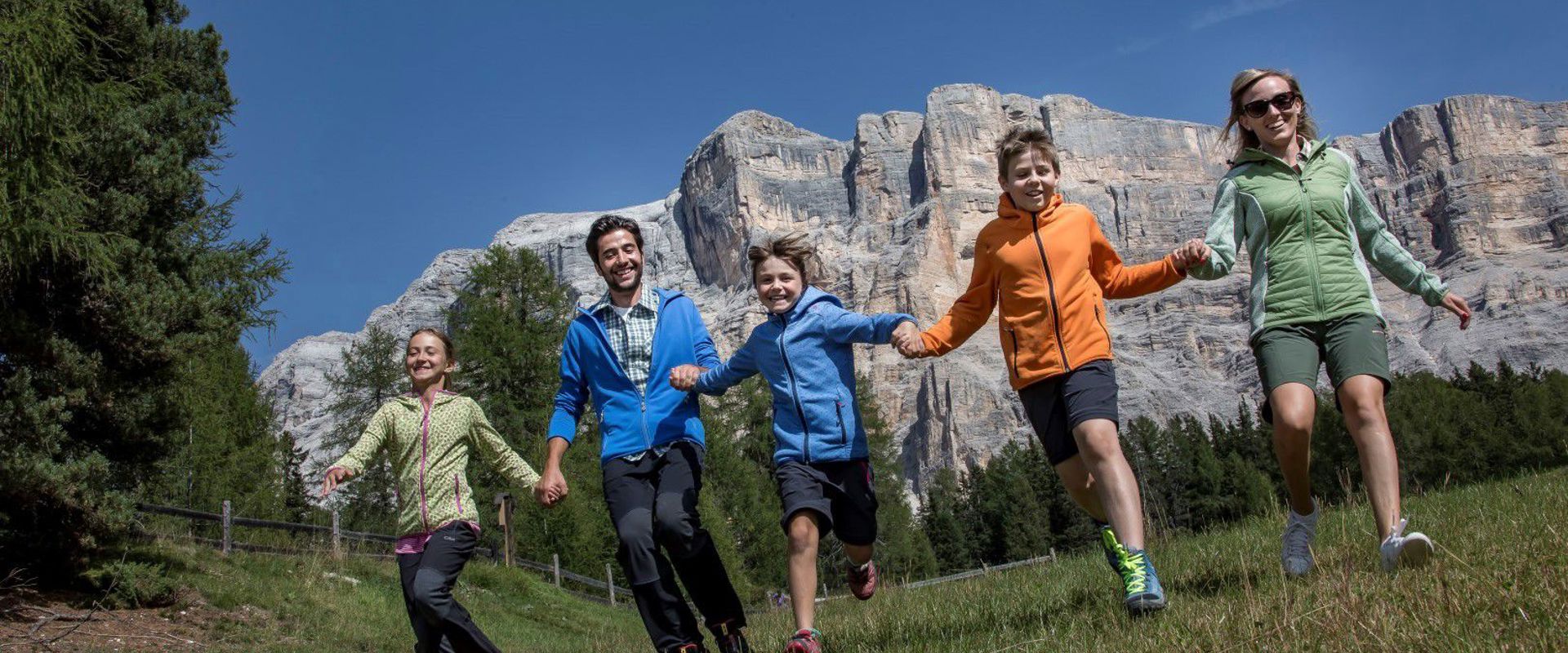 Famiglia corre su un prato tenendosi per mano. Sullo sfondo si intravedono ripide pareti rocciose.
