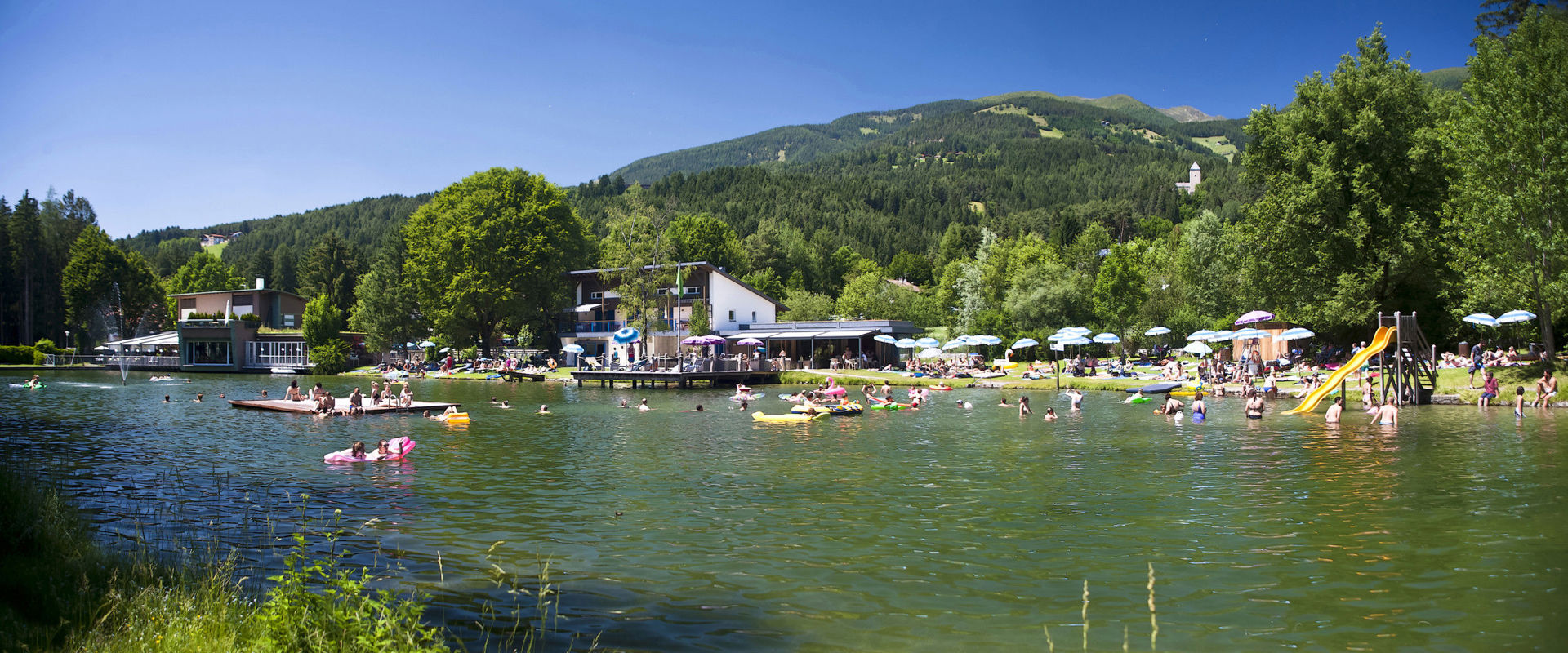 Lago balneabile con ristorante, pontile, zattera, scivolo e ombrelloni bianchi e blu.