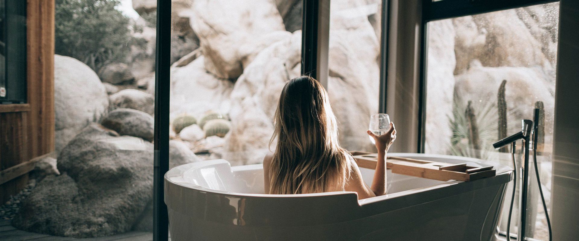 Davanti alla porta vetrata del patio una donna sta seduta nella vasca da bagno con in mano un bicchiere di vino bianco.