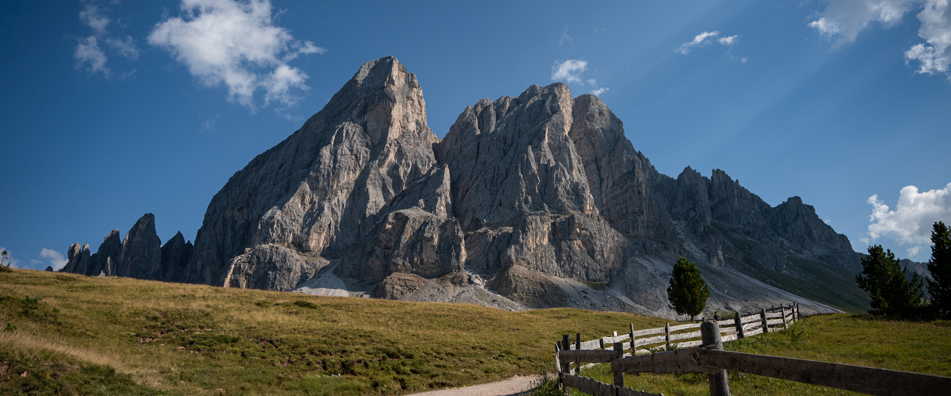 Un suggestivo massiccio montuoso si erge al Passo delle Erbe dietro un prato alpino con alberi sparsi e una recinzione in legno