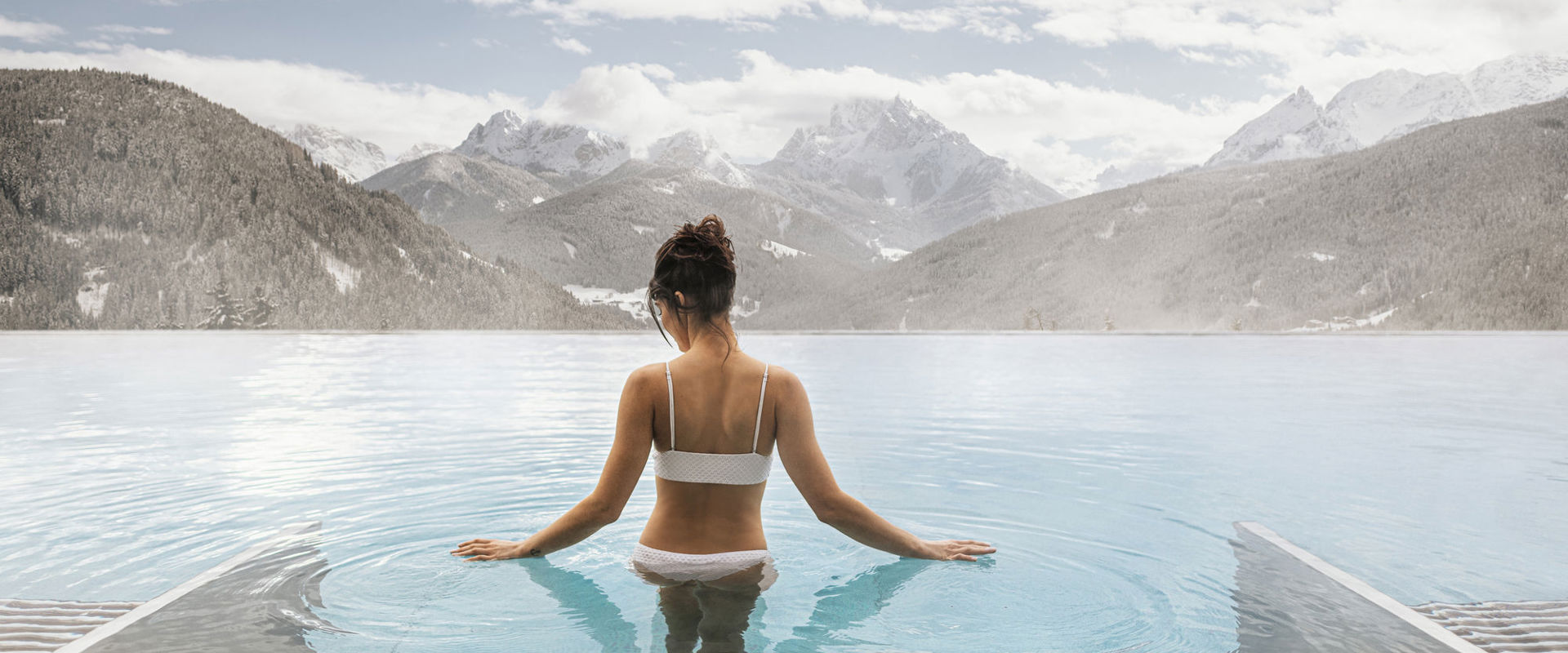 Piscina wellness all'aperto Donna in bikini bianco e capelli scuri legati, scende i gradini della piscina esterna fumante. Sullo sfondo si vedono le montagne innevate.