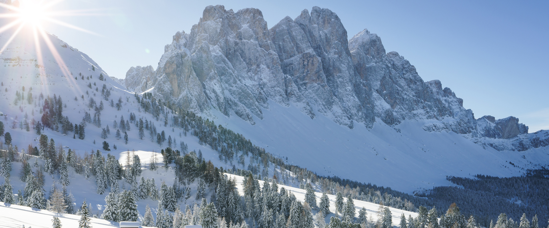 Funes Paesaggio montano soleggiato e innevato con rocce e alberi, davanti due scialpinisti in discesa.