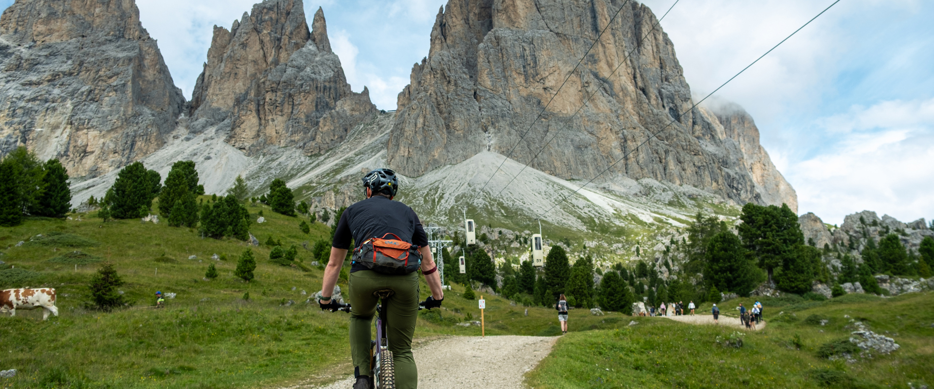 Ciclisti su sentiero sotto la funivia e davanti alle Dolomiti