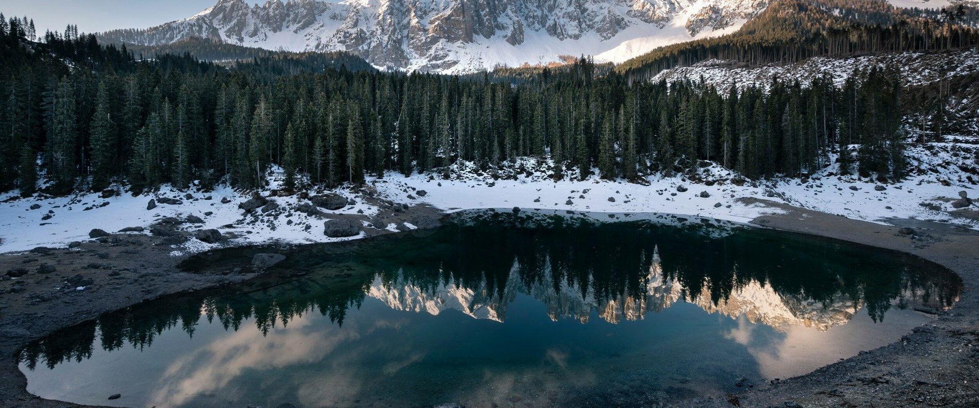 Lago di Carezza in inverno Paesaggio montano con rocce e conifere innevate, in parte illuminato dal sole e in parte in ombra, riflesso in un piccolo lago